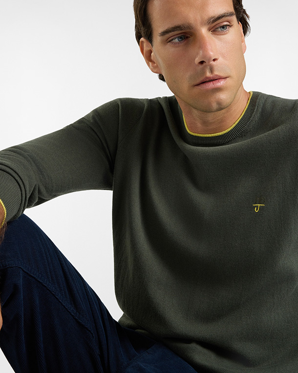 For the HB Jeans knitwear category, a model wears a dark olive green long-sleeved crewneck sweater featuring a subtle contrasting yellow trim on the collar and cuffs, embellished with a small yellow embroidered logo on the chest and paired with dark trousers against a light background.