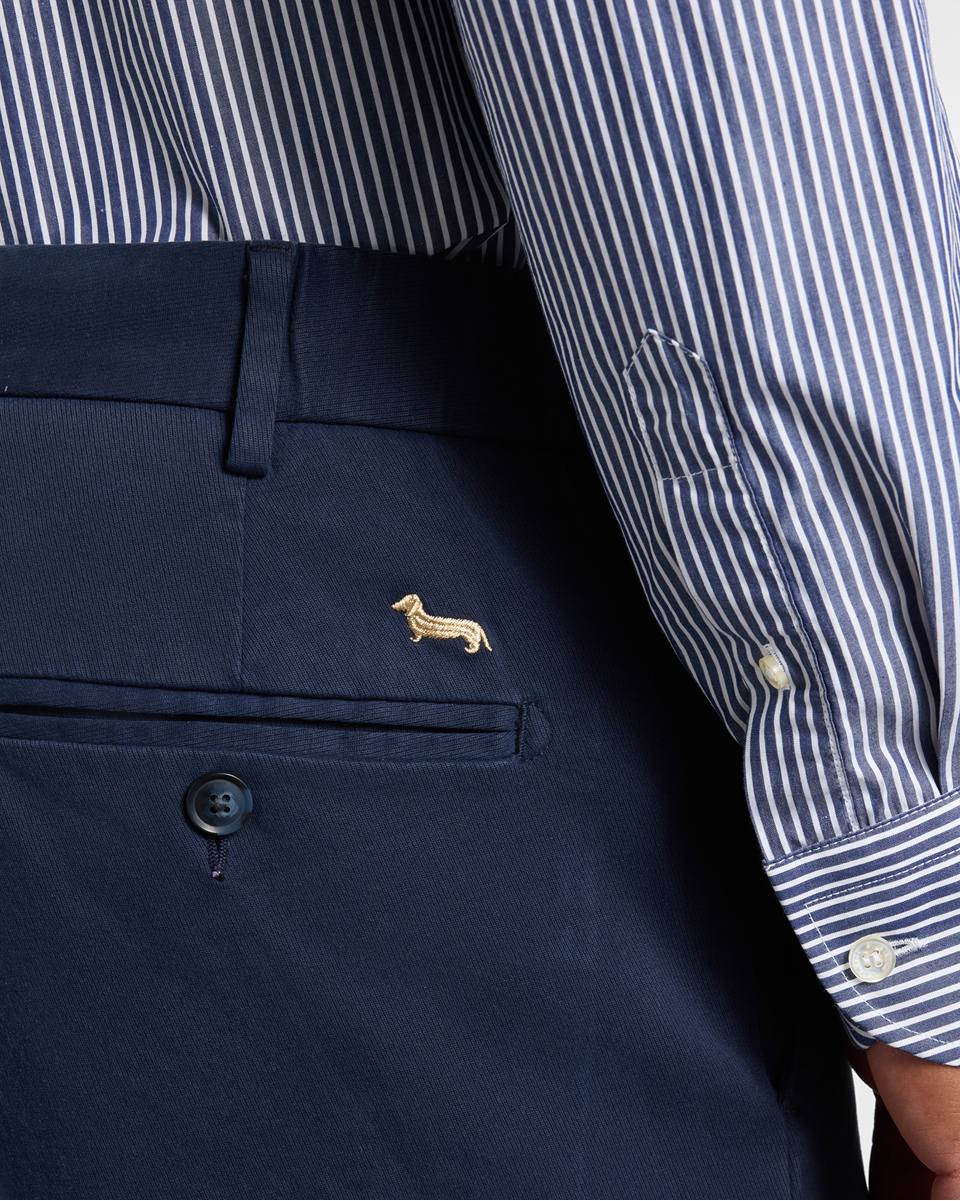 Dettaglio di pantaloni blu scuro con il logo del bassotto ricamato in oro sopra la tasca posteriore, abbinati a una camicia a righe bianche e blu.