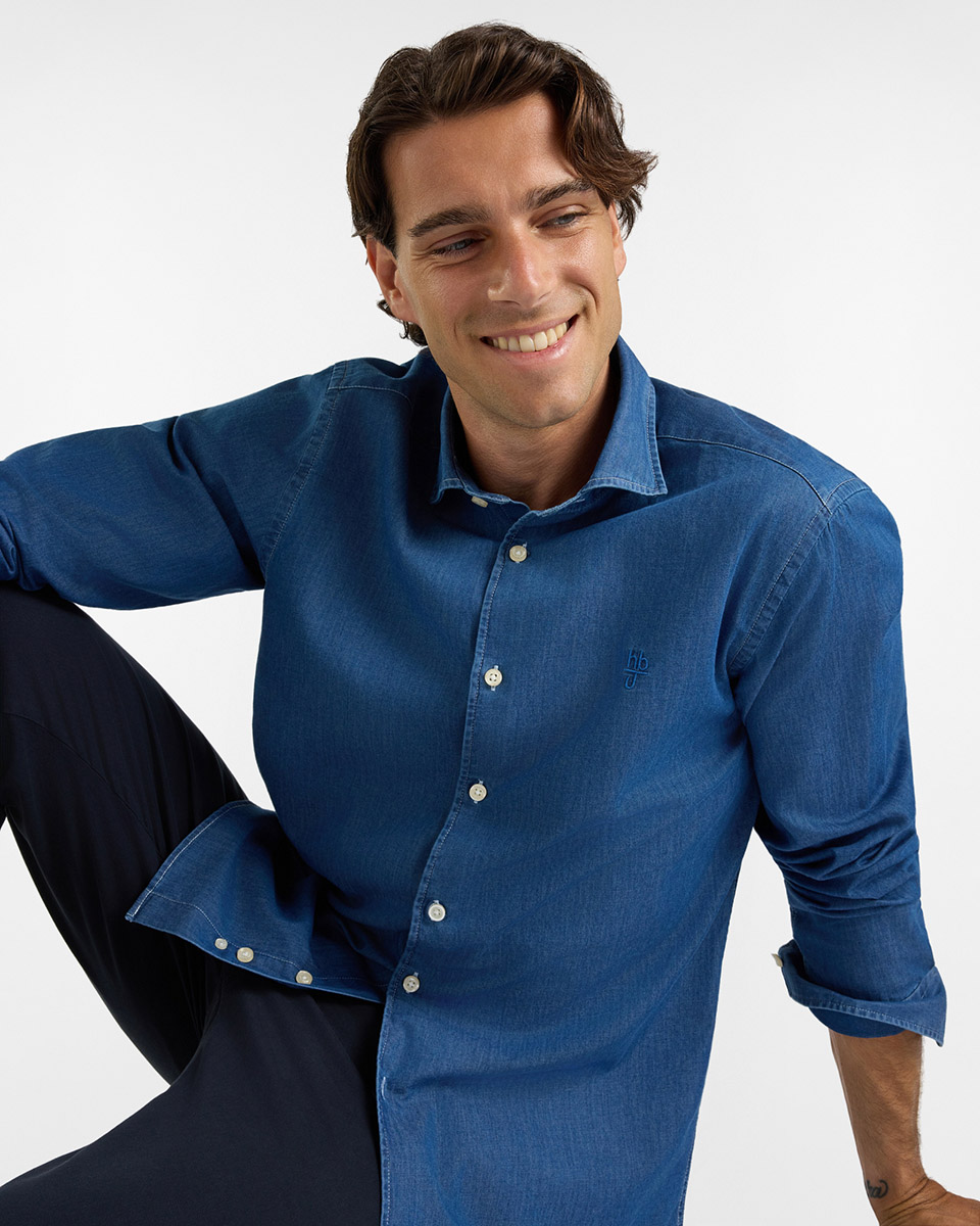 For the HB Jeans shirts category, a smiling model wears a medium-wash denim shirt with long, rolled-up sleeves, featuring a spread collar, contrasting light buttons, and a tone-on-tone embroidered logo on the chest, paired with dark trousers against a white background.
