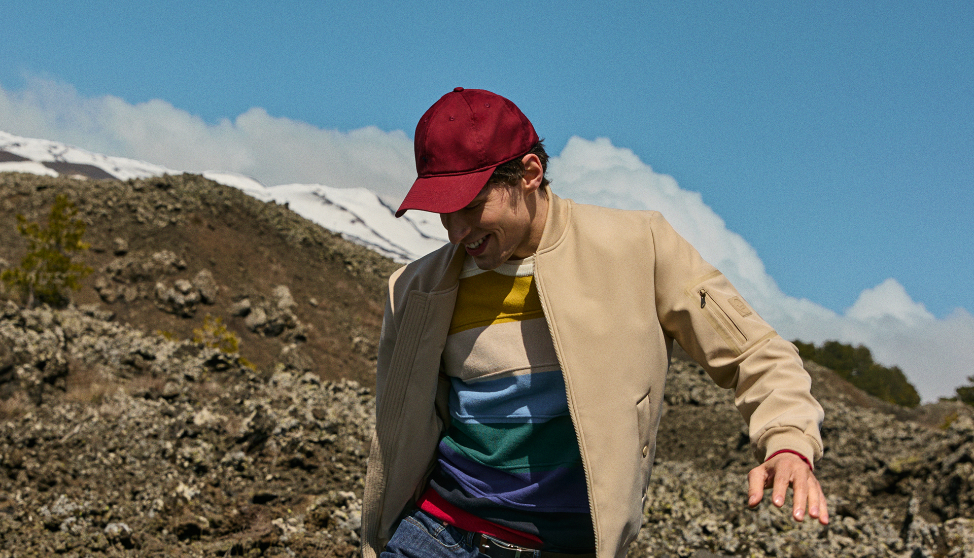 Un uomo con cappellino da baseball rosso bordeaux e giubbotto bomber beige aperto sopra una maglia a righe colorate sorride in un paesaggio vulcanico con montagne innevate sullo sfondo.