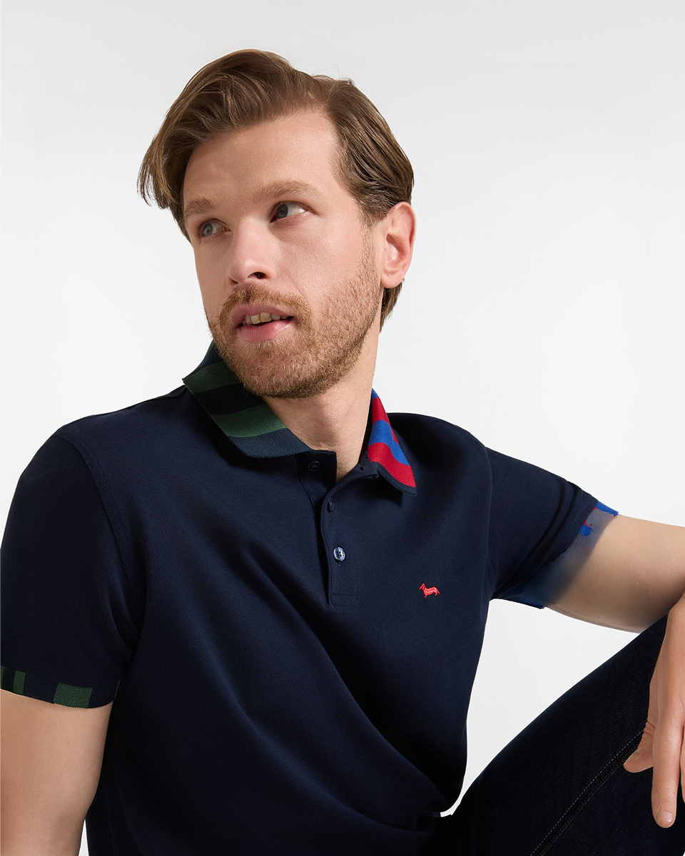 A half-length photograph, part of the FW26 campaign themed "Polo Shirts for Him", portrays a man with dark blonde hair and a beard, seated and turned in a three-quarter view to the left. He is wearing a dark blue polo shirt with dark buttons, a collar decorated with dark green, dark red, and purple colored stripes, and the brand's logo embroidered in red on the chest. The background is a solid white.