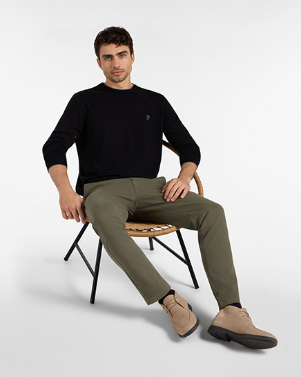 For the HB Jeans trousers category, a model sits on a chair wearing olive green chino trousers, a black long-sleeved sweater, and beige suede desert boots against a white background.