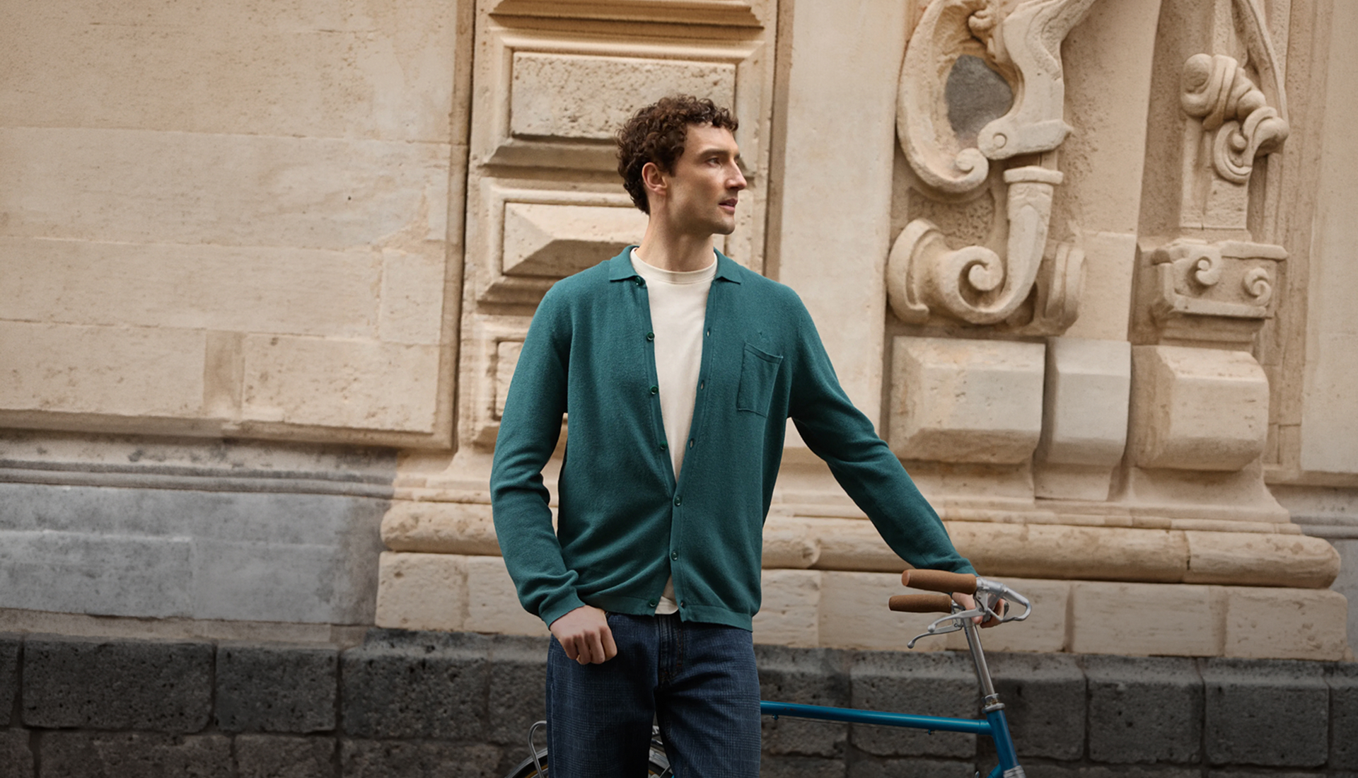 Un uomo con capelli ricci indossa un cardigan verde petrolio bottoni aperti su una t-shirt panna e jeans blu, accanto a una bicicletta d'epoca con manubrio in pelle, davanti a un edificio storico in pietra con dettagli barocchi.