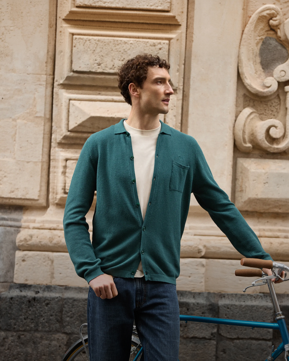 Un uomo con capelli ricci indossa un cardigan verde petrolio bottoni aperti su una t-shirt panna e jeans blu, accanto a una bicicletta d'epoca con manubrio in pelle, davanti a un edificio storico in pietra con dettagli barocchi.