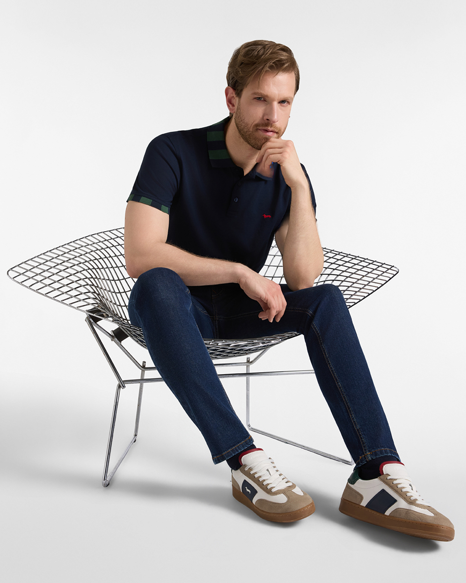 A blond man with a beard, sitting on a chair with a chromed steel wire mesh frame, wearing a navy blue short-sleeved polo shirt with green and navy blue striped collar and cuff trim, navy blue jeans and dark grey and beige sneakers.