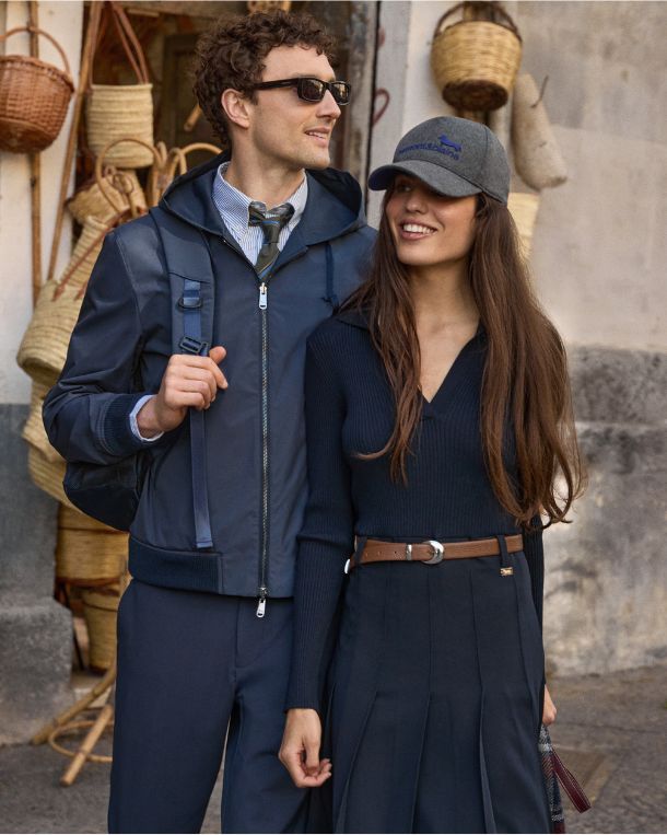 The image shows a smiling couple walking outside an artisan shop decorated with woven wicker baskets hanging on the walls. The man is wearing dark sunglasses, a dark blue hooded windbreaker over a striped shirt and tie, matching trousers, and a backpack over his shoulder. The woman beside him is wearing a dark blue long-sleeved ribbed top with a V-neck, a matching pleated skirt with a brown leather belt, and a grey baseball cap featuring a blue embroidered logo.