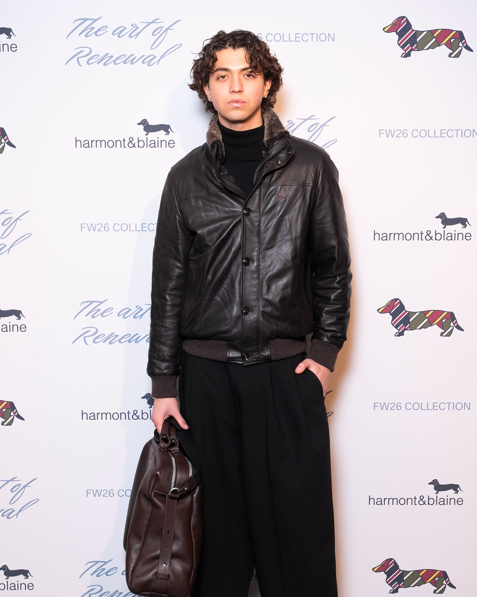 Man wearing black leather jacket and wide dark trousers holding a bag
