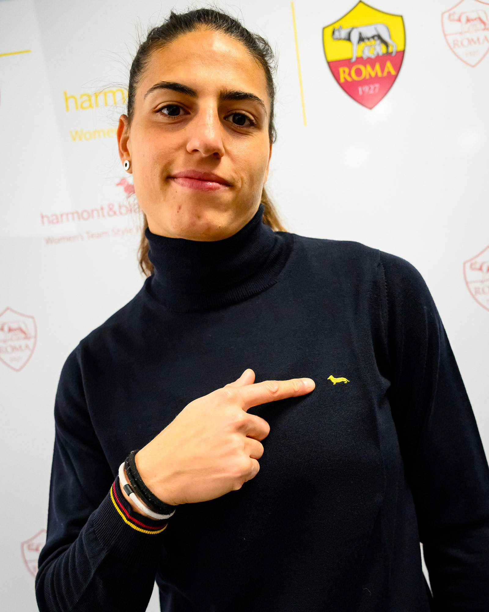 Woman wearing a navy blue turtleneck pointing at the yellow logo.