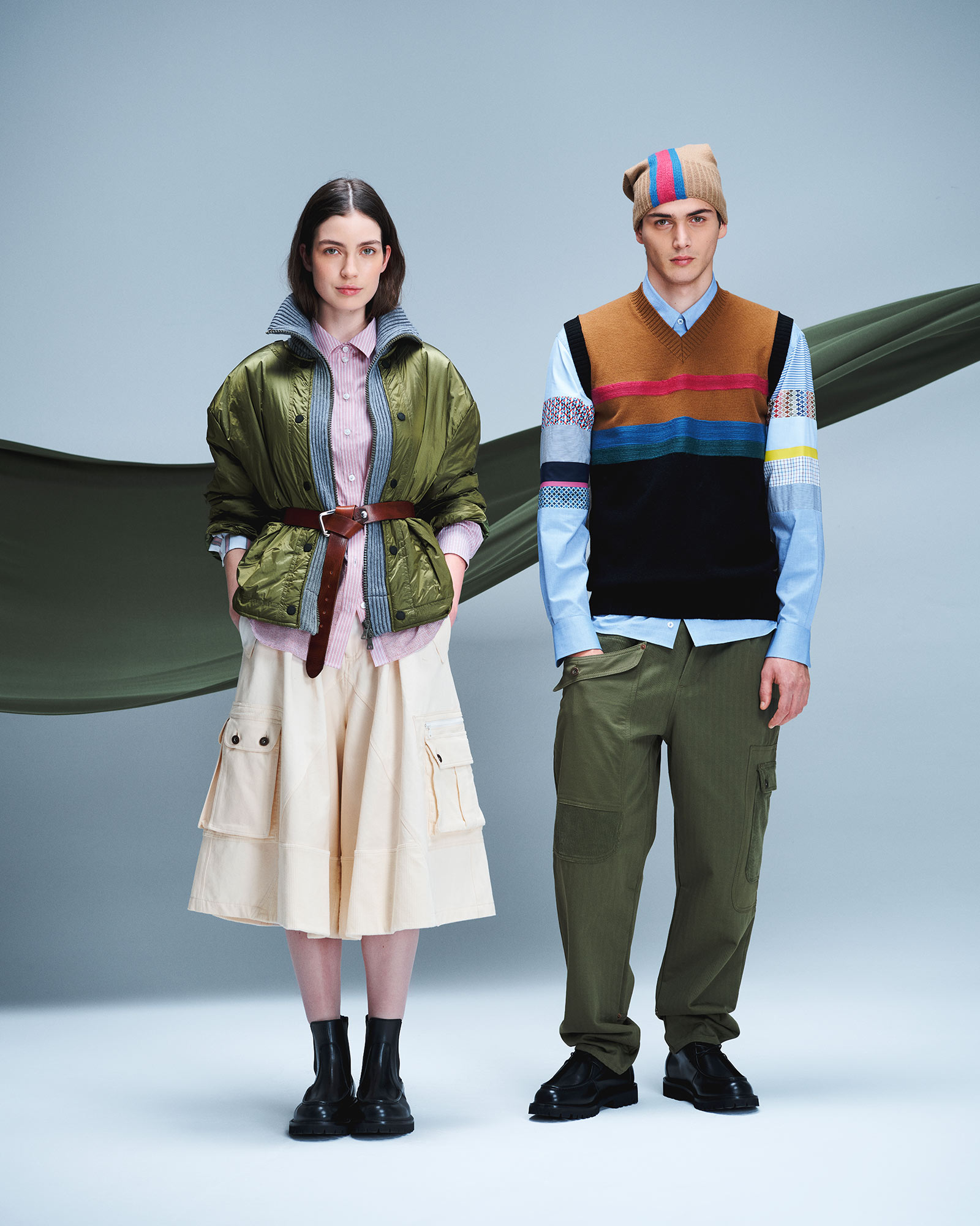 Woman and man in utility looks; she wears green bomber jacket and light cargo skirt, he wears multicolor vest and green cargo trousers