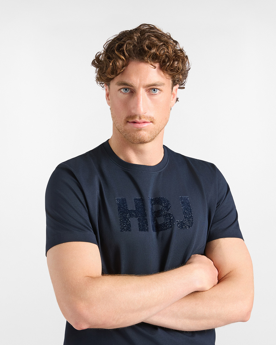 For the HB Jeans t-shirts category, a model with crossed arms wears a dark blue short-sleeved crewneck t-shirt featuring a tone-on-tone textured "HBJ" logo printed across the chest against a white background.